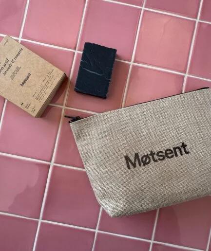 Trousse de toilette écoresponsable Motsent en toile de jute, accompagnée d’un savon au charbon actif bio pour le visage et le corps, posée sur un carrelage rose.