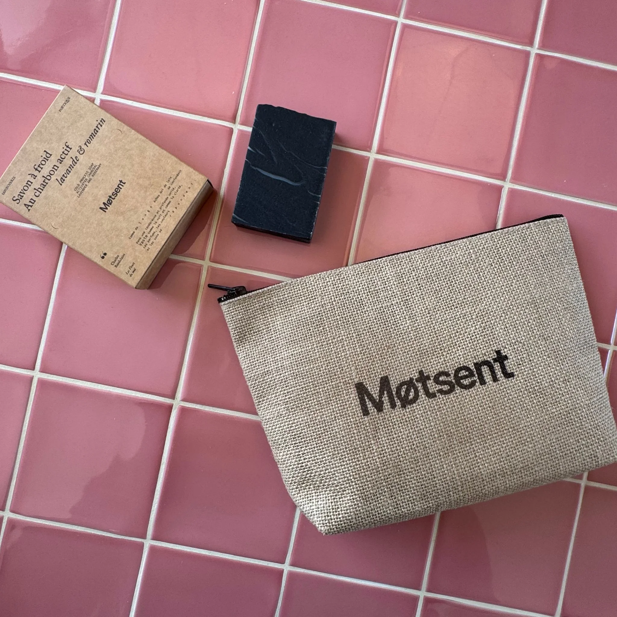 Trousse de toilette éthique Motsent avec savon au charbon actif bio – fabriquée en France Trousse de toilette écoresponsable Motsent en toile de jute, accompagnée d’un savon au charbon actif bio pour le visage et le corps, posée sur un carrelage rose.